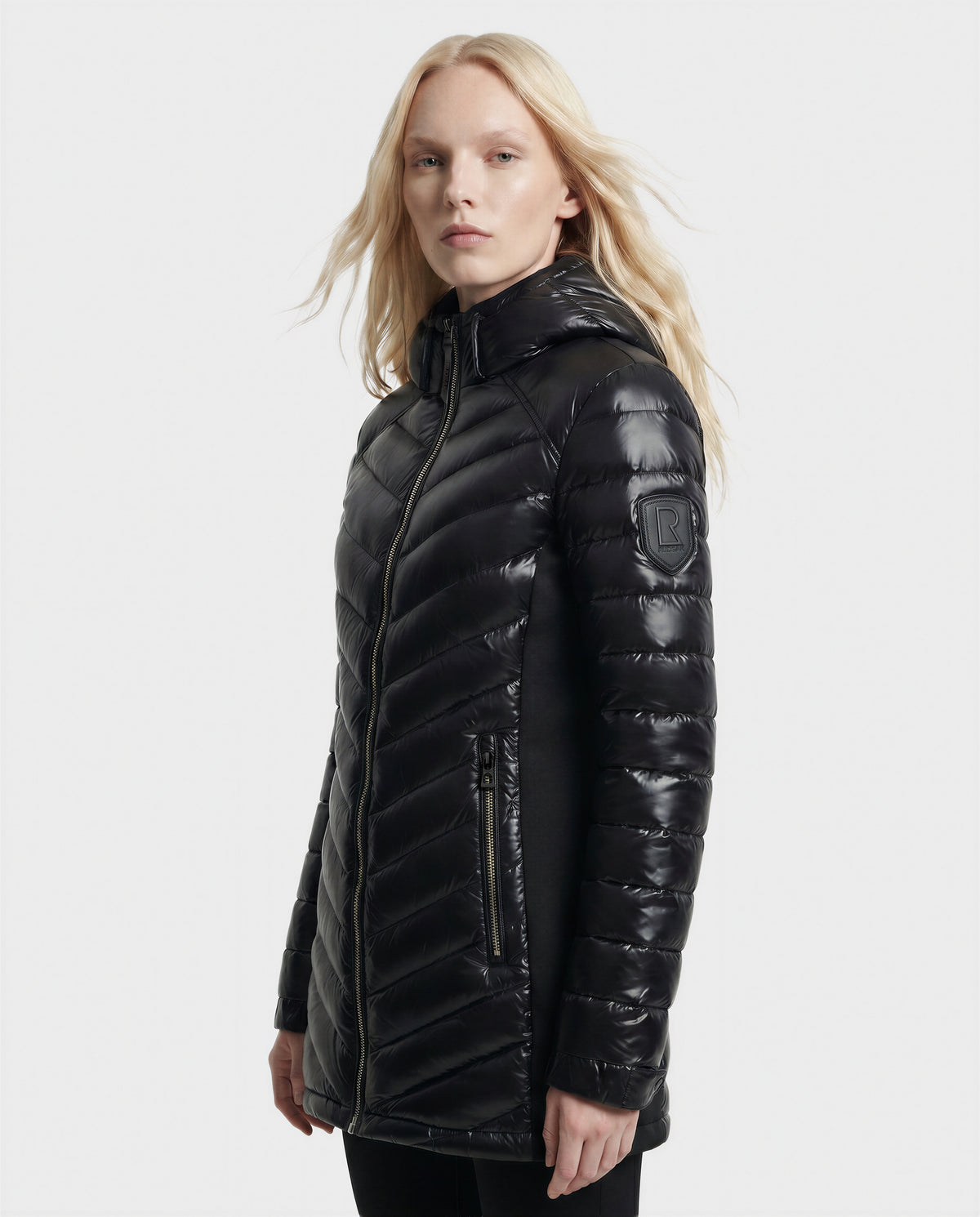 A person with long blonde hair wears the Rudsak COSETTE C - L124011 Black women's fitted down jacket in shiny black with a hood, quilted design, sleeve patch, and zippered pockets, standing against a plain light background.