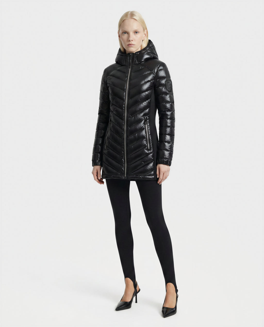 A woman stands against a plain white background, wearing the Rudsak COSETTE C - L124011 Black fitted down jacket with a hood, paired with black leggings and high-heeled shoes. She faces forward with her arms by her sides.