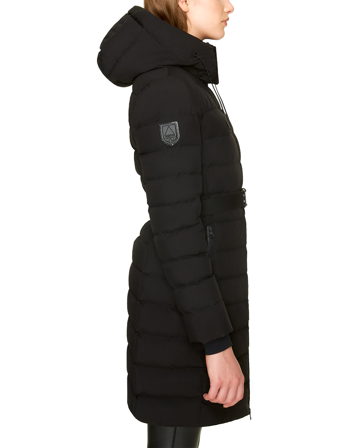 A woman in profile wears the Rudsak SAMIRA - 4124503 Black women's quilted down puffer, featuring a patch on the upper sleeve, belted waist, removable hood, and premium insulation. She pairs it with black pants and has straight light brown hair.