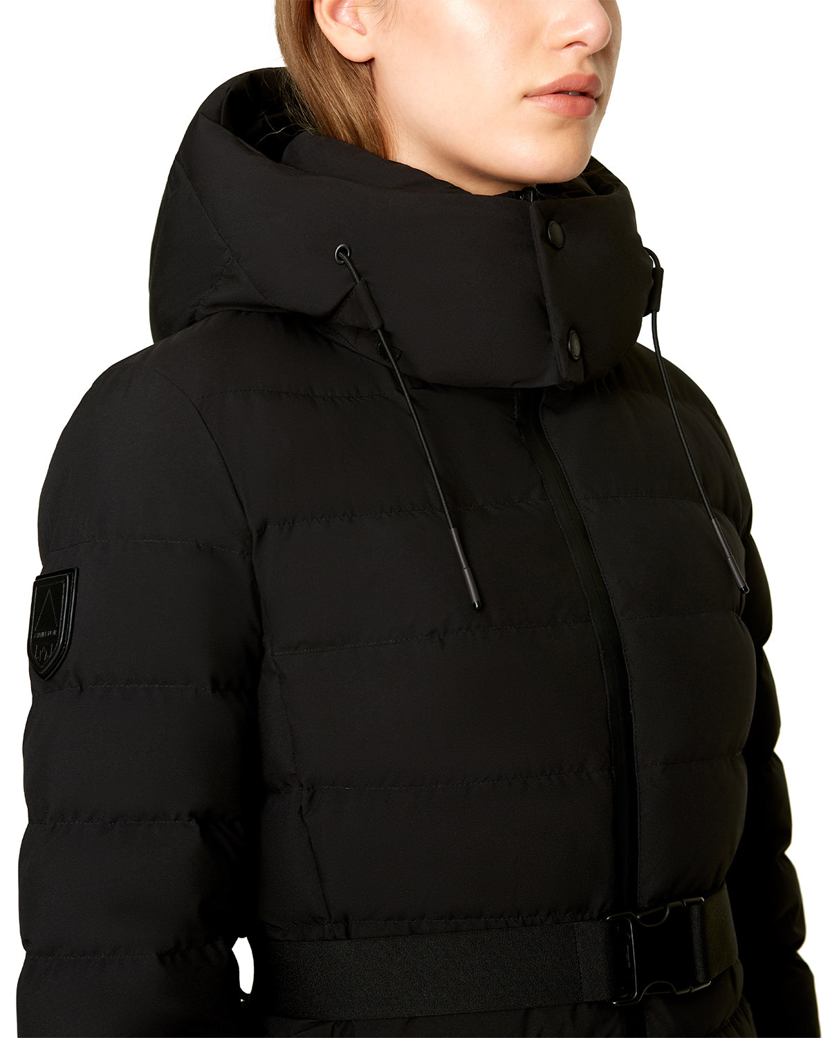 A person wearing the Rudsak SAMIRA - 4124503 Black women's quilted down puffer with a high hood, drawstrings, and black belt. The photo is cropped to show part of the upper chest and face without visible expression.