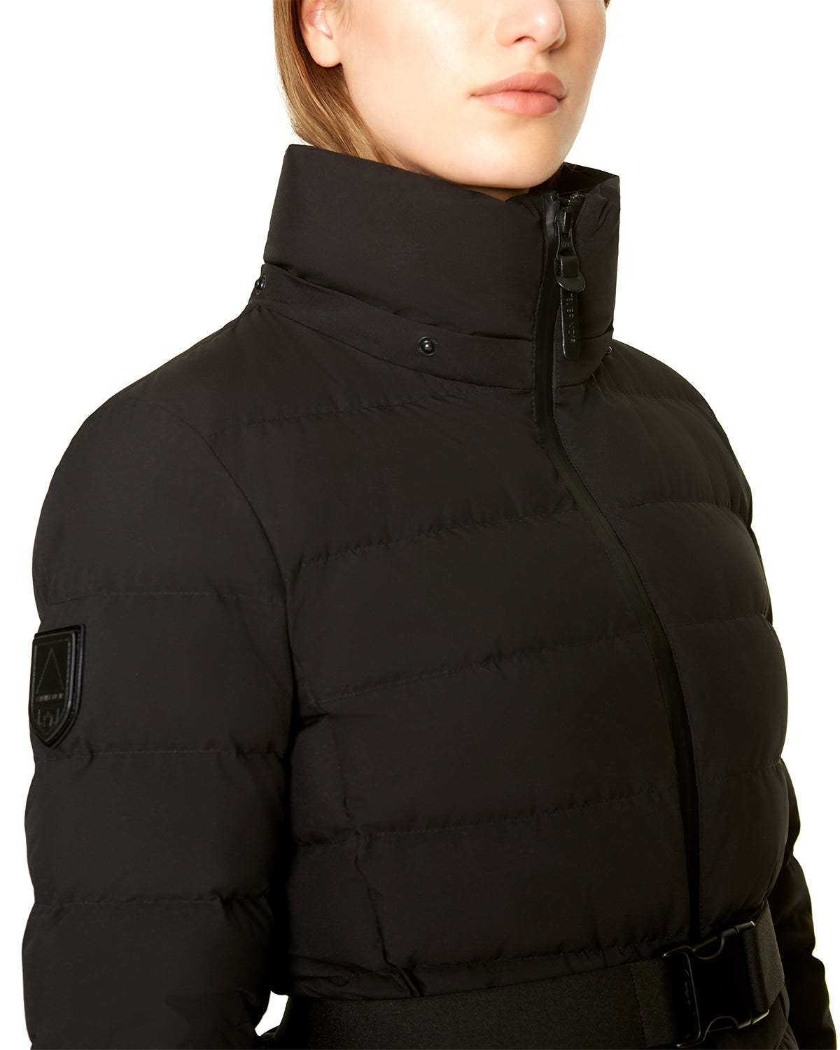 Wearing the Rudsak SAMIRA - 4124503 Black women's quilted down puffer with high collar, zipper, belt, removable hood, and a patch on the upper arm. Only the person's lower face and shoulders are visible.