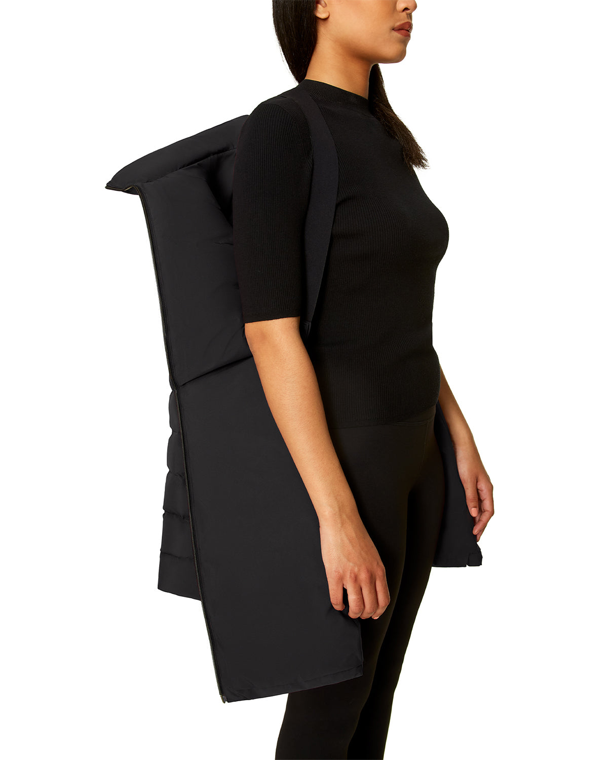A woman in a black short-sleeve top and pants is shown from the side, wearing the Rudsak SAMIRA - 4124503 Black—a large, padded item with shoulder straps resembling an oversized quilted down puffer or wearable pillow.