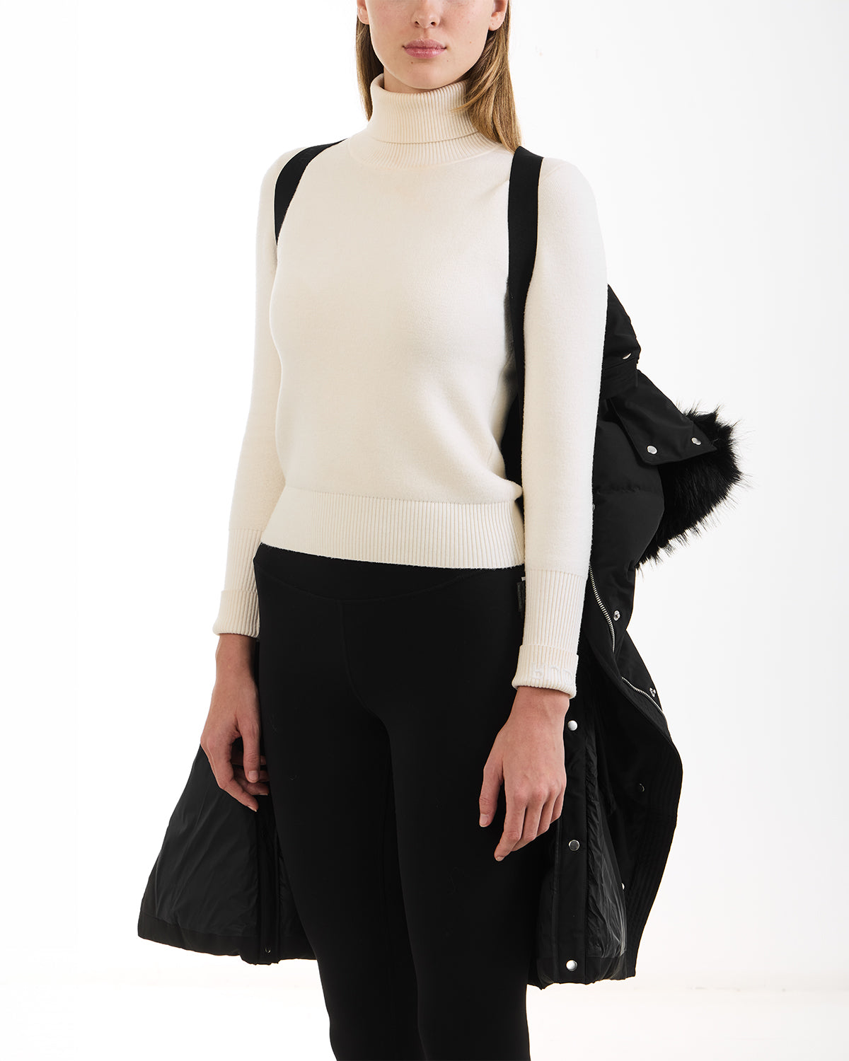 A woman stands against a white background, draping the Rudsak JANET - 4125504 Black coat with premium down over her arm. She wears a white turtleneck sweater and black pants, her face partially visible.