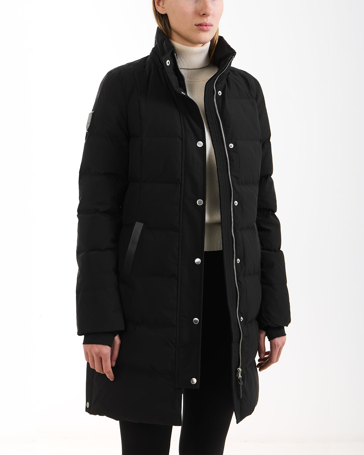 A person wearing the Rudsak JANET - 4125504 Black long quilted down puffer jacket with snap buttons, high collar, side pockets, and premium insulation over a light turtleneck, standing against a white background.
