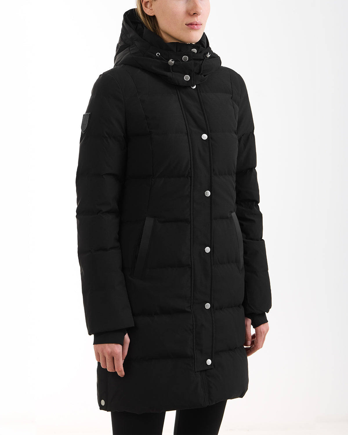 A person wears the Rudsak JANET - 4125504 Black long quilted down puffer jacket with a hood, side pockets, snap buttons, and an upper sleeve patch. The face is partially visible against a plain white background.