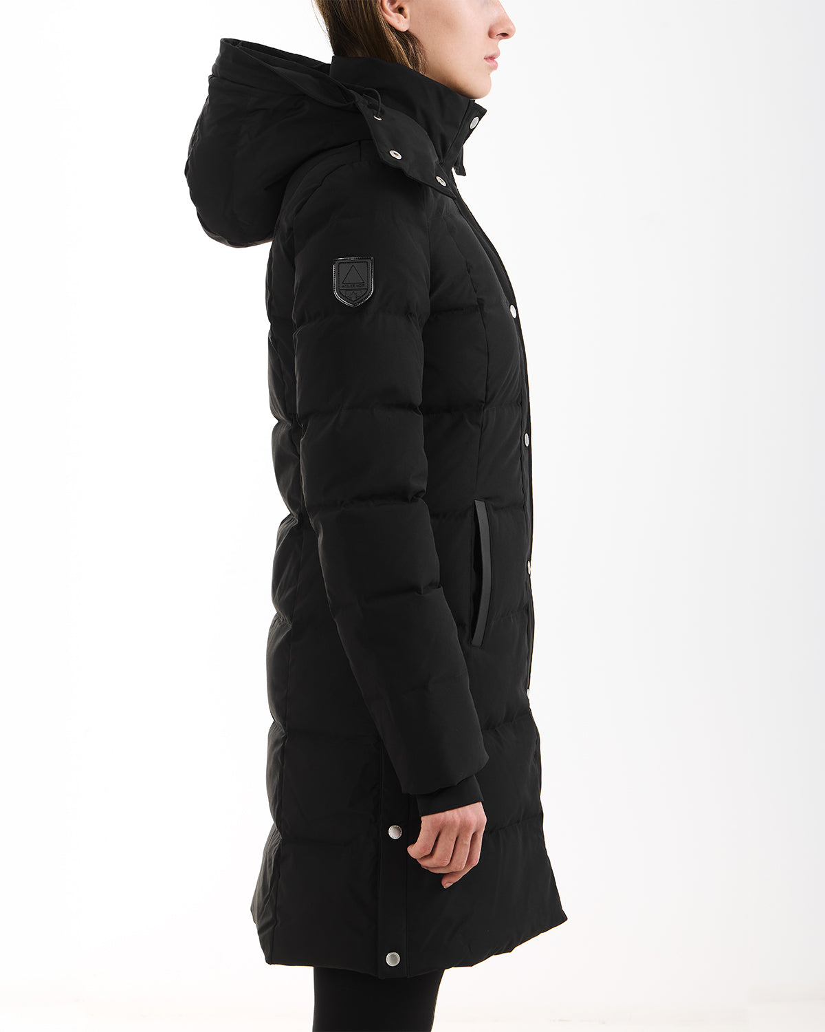 A person wearing the Rudsak JANET - 4125504 Black, a black, hooded knee-length down puffer jacket with snap buttons, an upper sleeve patch, and premium down insulation stands in profile against a white background.