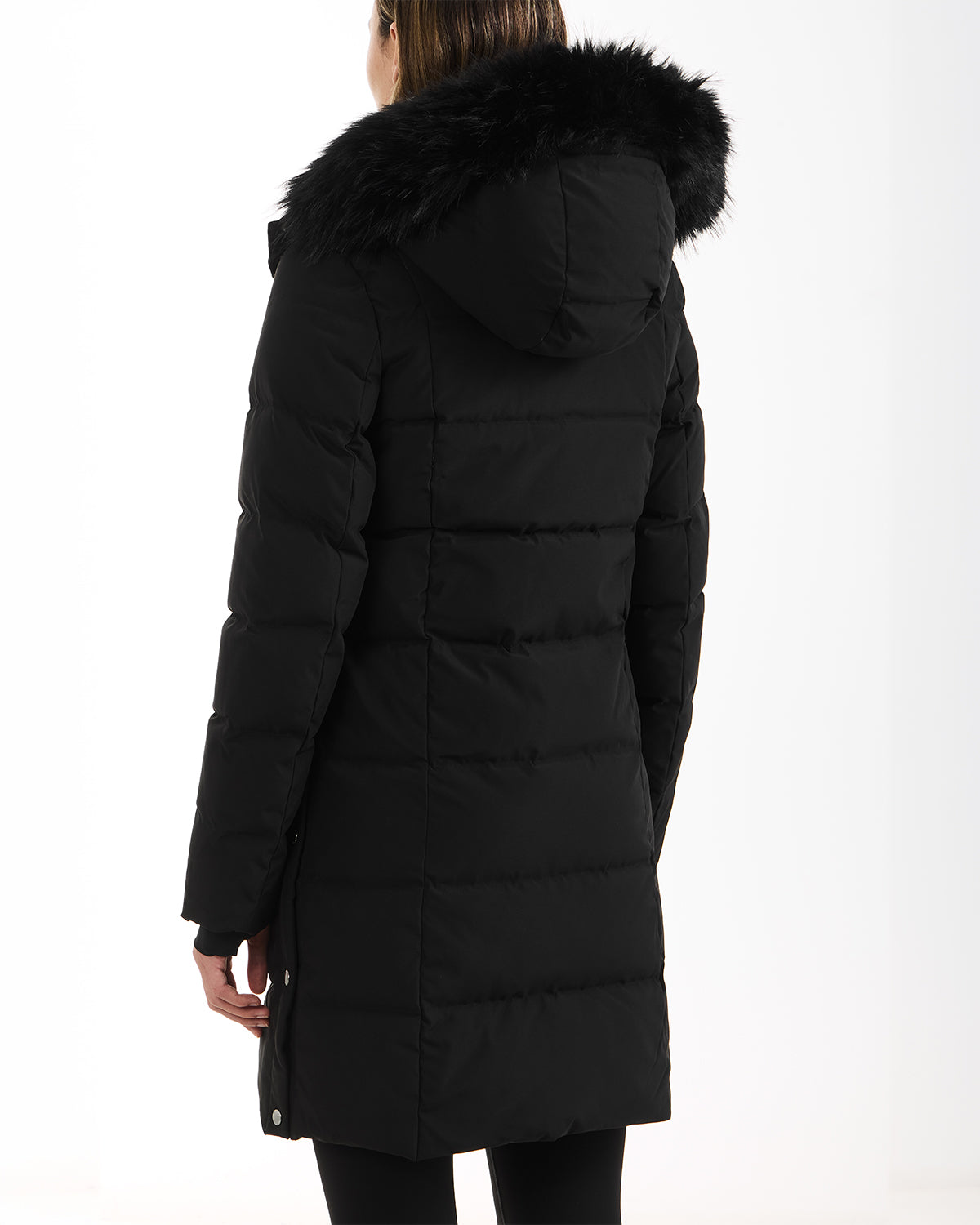 A woman with straight brown hair is shown from the back wearing the Rudsak JANET - 4125504 Black long quilted down puffer jacket with a black faux fur-trimmed hood, standing against a plain white background.