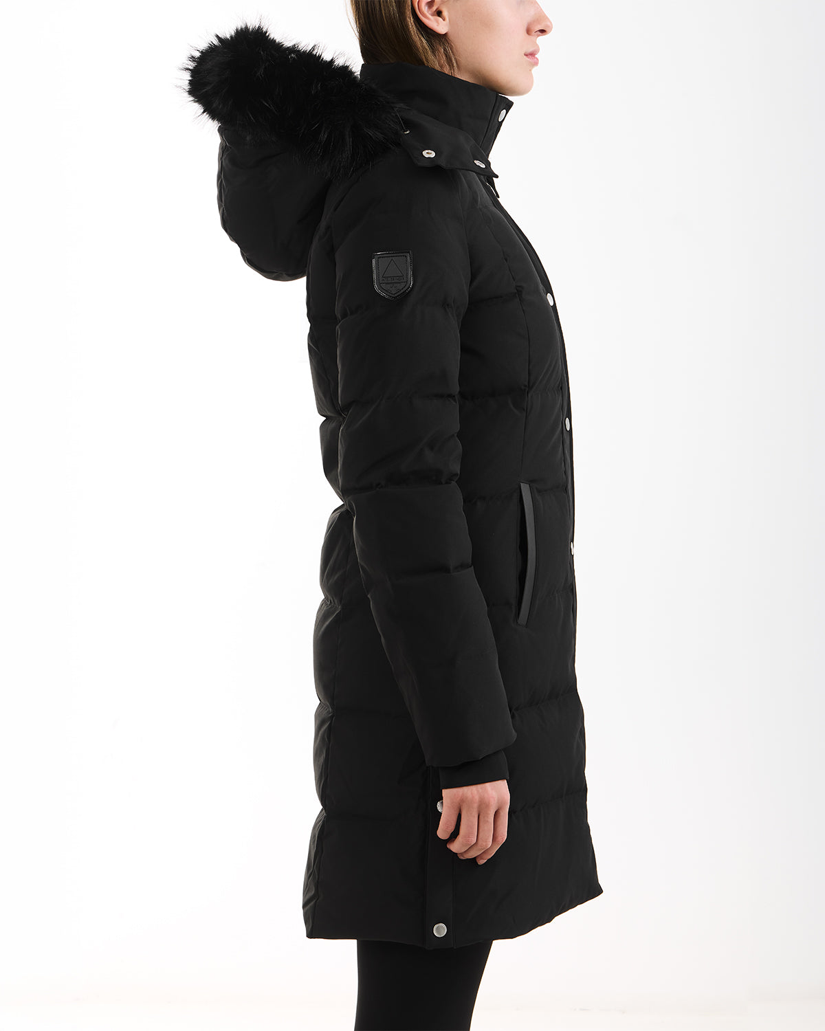 A person stands in profile against a white background wearing the Rudsak JANET - 4125504 Black, a long black quilted down puffer jacket with hooded black fur trim, premium down insulation, snap-button front, and upper arm patch.