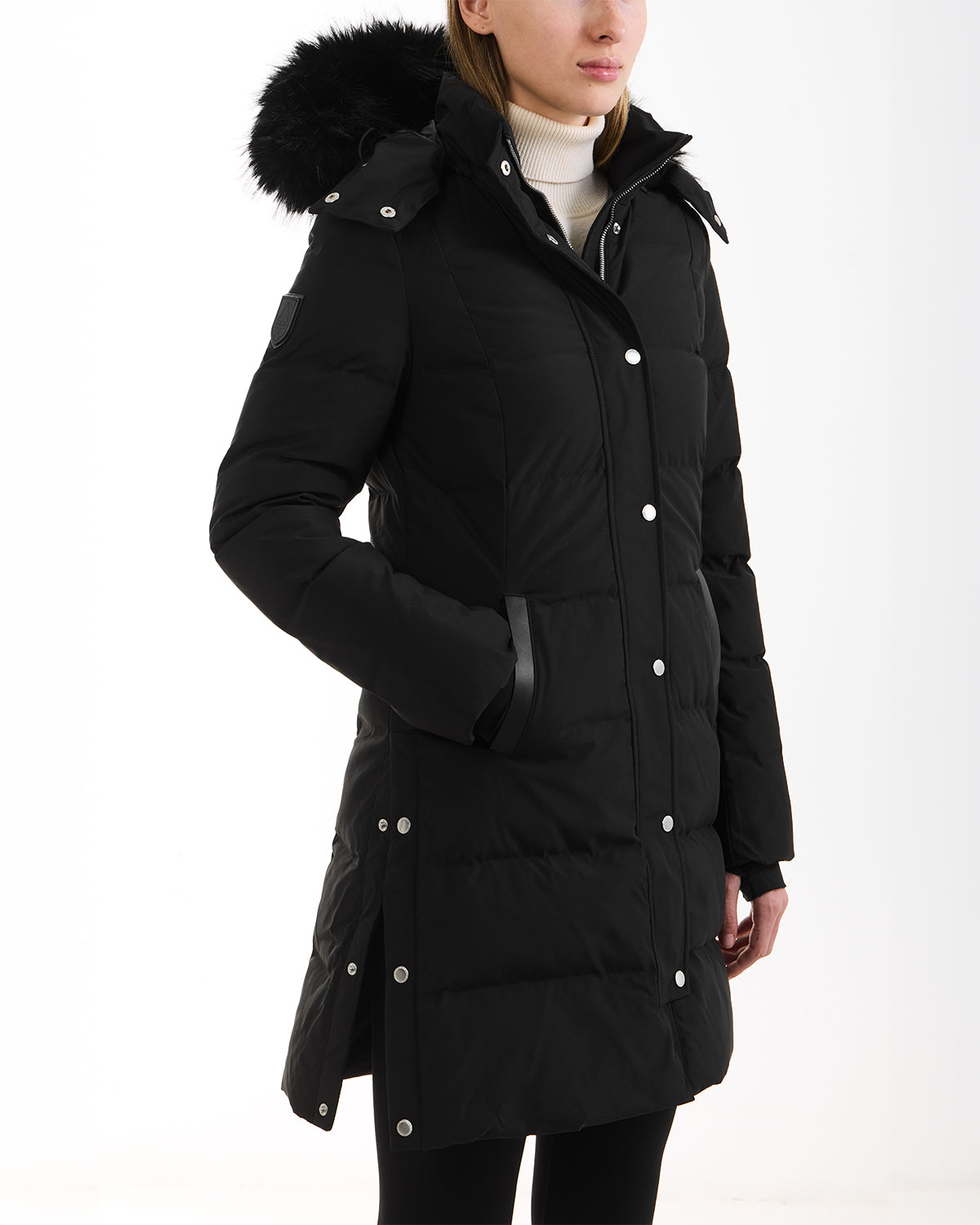 A woman wears the Rudsak JANET - 4125504 Black long quilted down puffer jacket with a removable faux fur collar, styled with black pants and a light turtleneck sweater, standing against a plain white background.