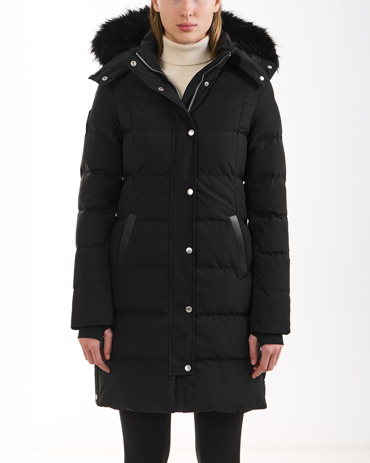 A woman models the Rudsak JANET - 4125504 Black long quilted down puffer jacket with a removable faux fur collar over a cream turtleneck, standing against a plain white background with her hands by her sides.