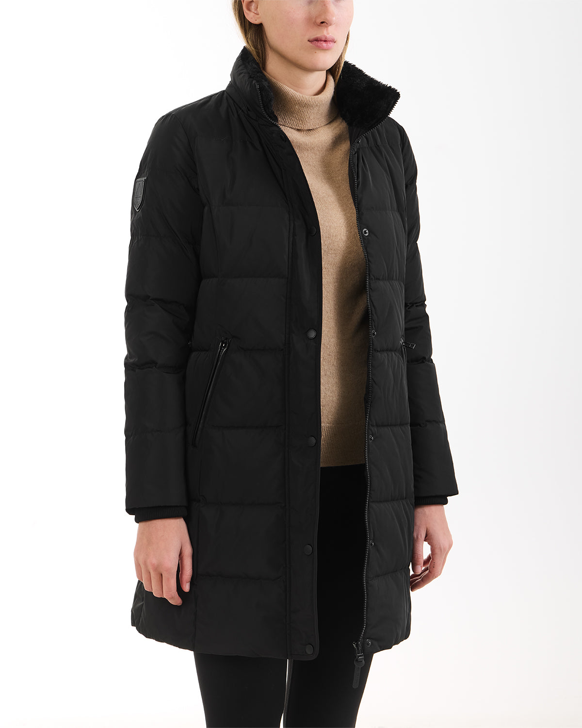 A woman wearing the Rudsak JANET - 4125504 Black knee-length quilted down puffer jacket over a beige turtleneck sweater and black pants, standing against a plain white background.