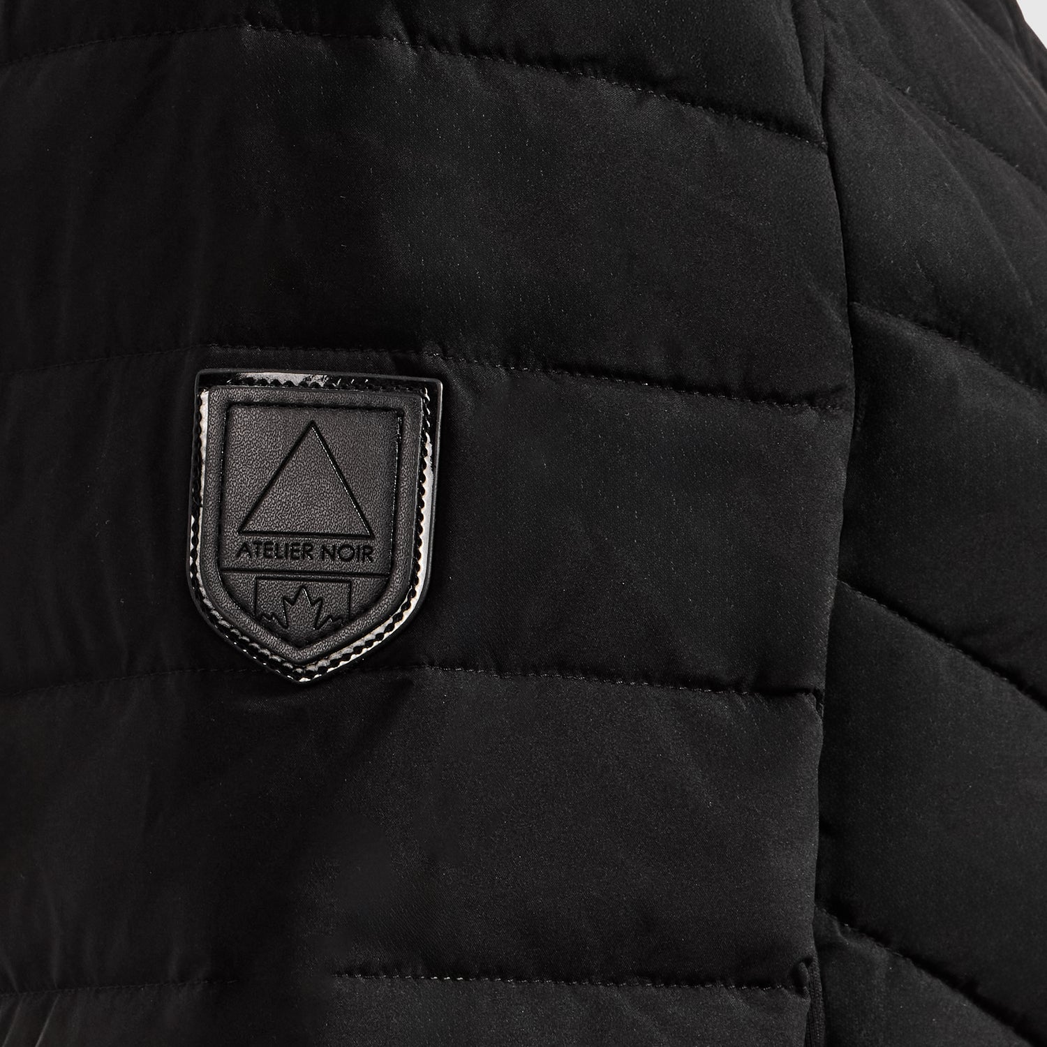 Close-up of the CORDELIA - 4124519 Black quilted down puffer jacket by Rudsak, featuring a patch with a triangle, "ATELIER NOIR" text, and a small maple leaf at the bottom.