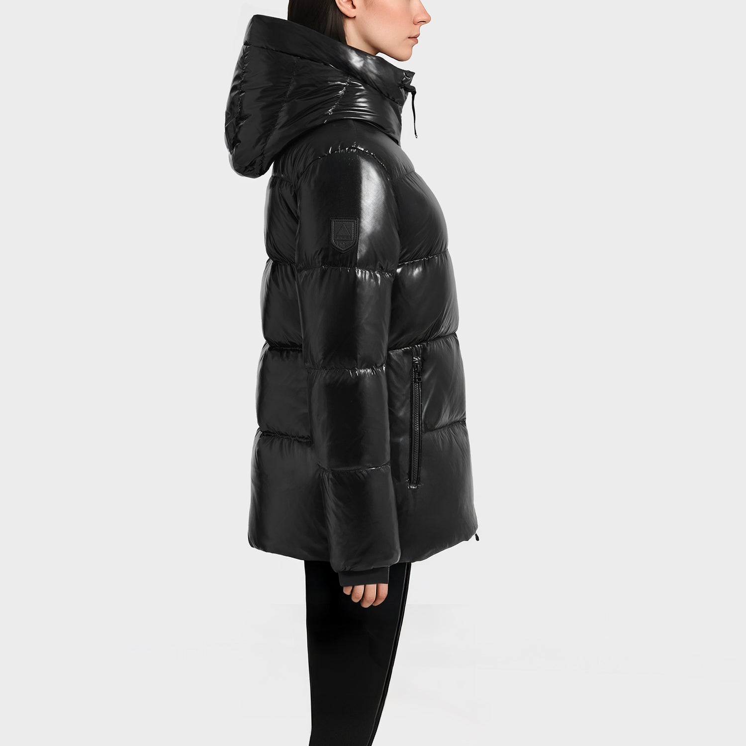 A person in profile wears the Rudsak MARY - 4124500 Black, a shiny black, hooded, mid-thigh puffer jacket with a quilted pattern and water-repellent fabric, standing against a plain light gray background.