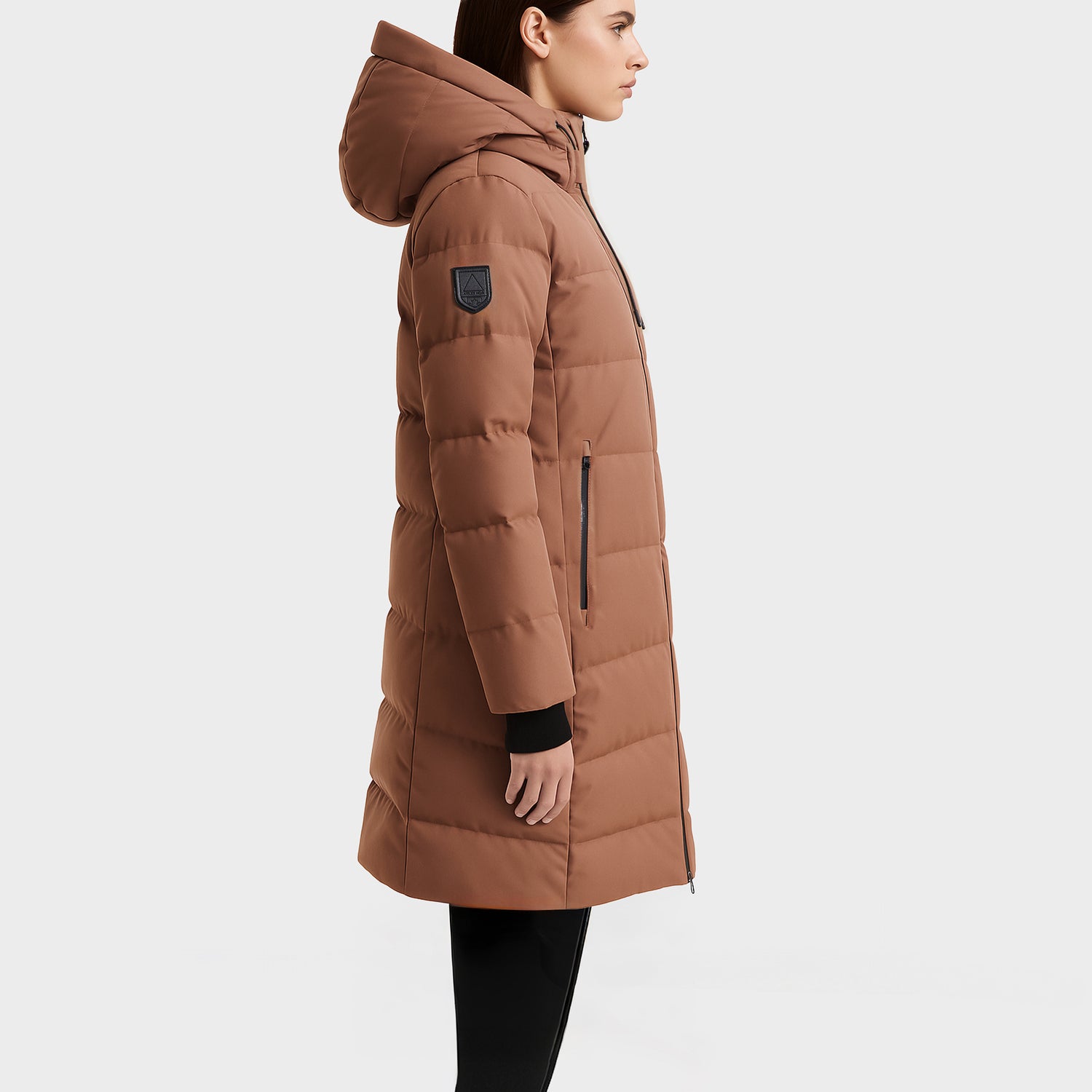 A person in profile wears the Rudsak SYDNEY COM - 4123511 Taupe, a long taupe water-repellent puffer coat with a hood, black zipper pockets, and an upper sleeve patch against a plain white background.