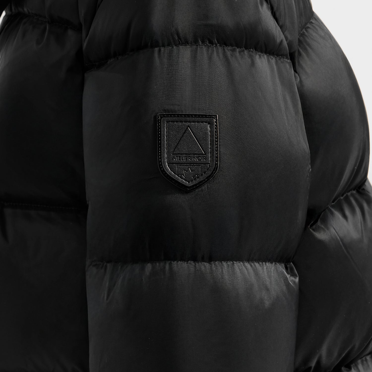 Close-up of the OLIVIA - 4123509 Black puffer jacket by Rudsak, featuring a shiny, quilted texture and a stitched patch logo with a triangle and "Alersindir" on the chest.