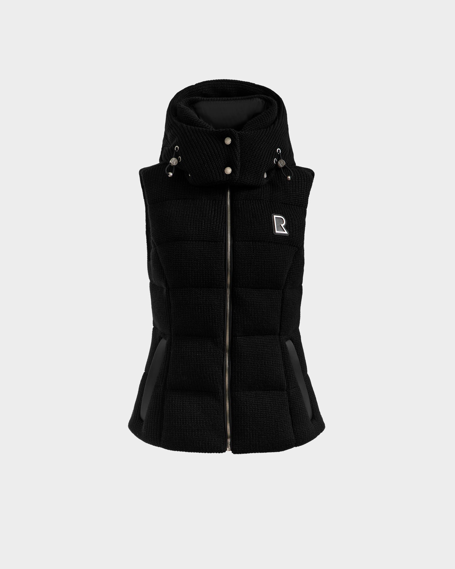 The Rudsak CASSIDY - 8125622 Black is a women’s black quilted down vest with a high buttoned collar, two front pockets, and a silver R logo on the left chest, shown against a plain light gray background.