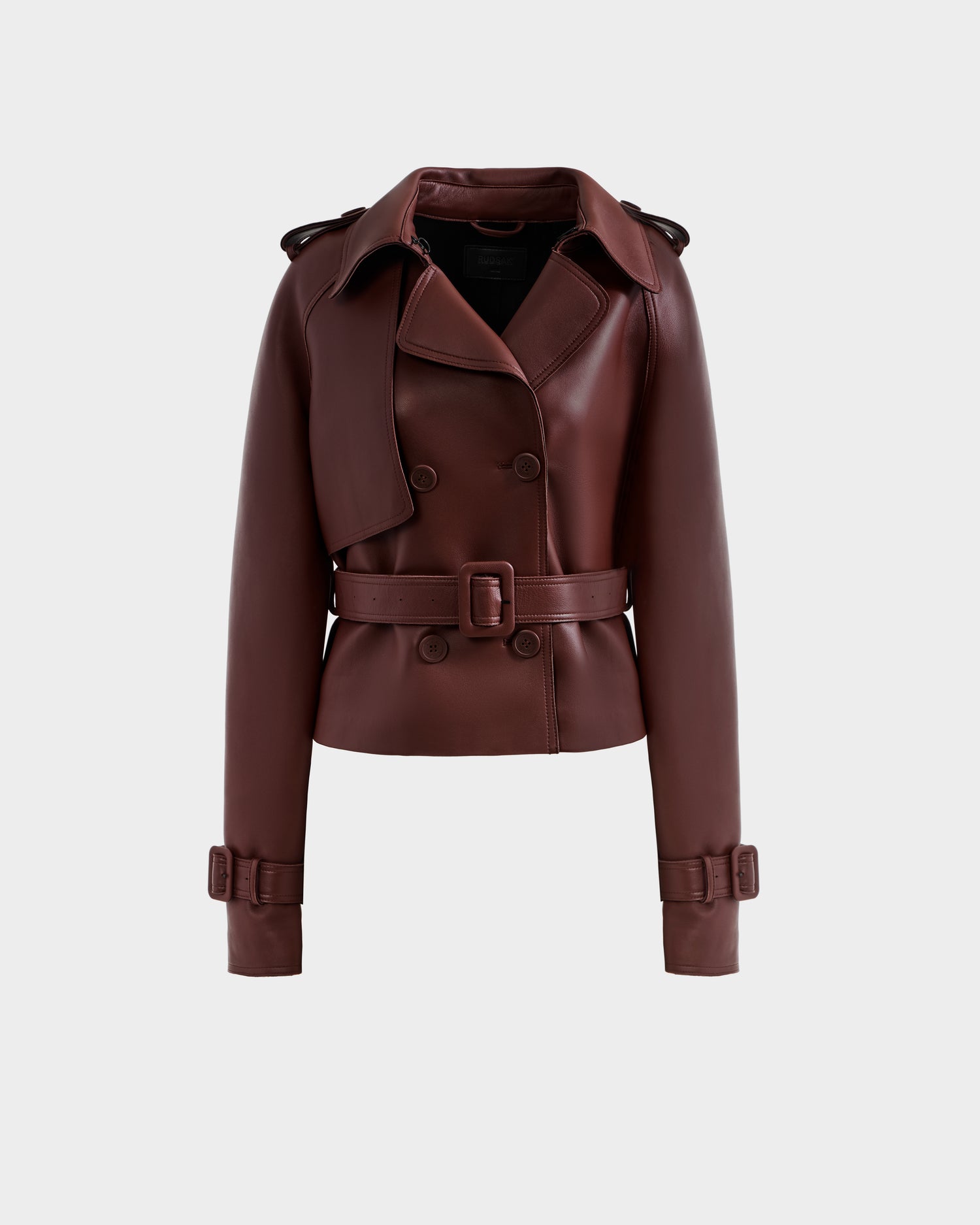 The CASSIE S-B - 8125554 Rust by Rudsak is a dark brown bonded leather cropped trench coat with a wide lapel, double-breasted buttons, belted waist, and buckled straps at the cuffs, shown against a plain light gray background.