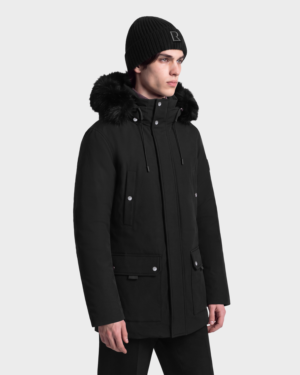 A man wearing an EDWARD - 6126501 Black deep-winter parka with a fur-trimmed hood and a matching knit beanie.