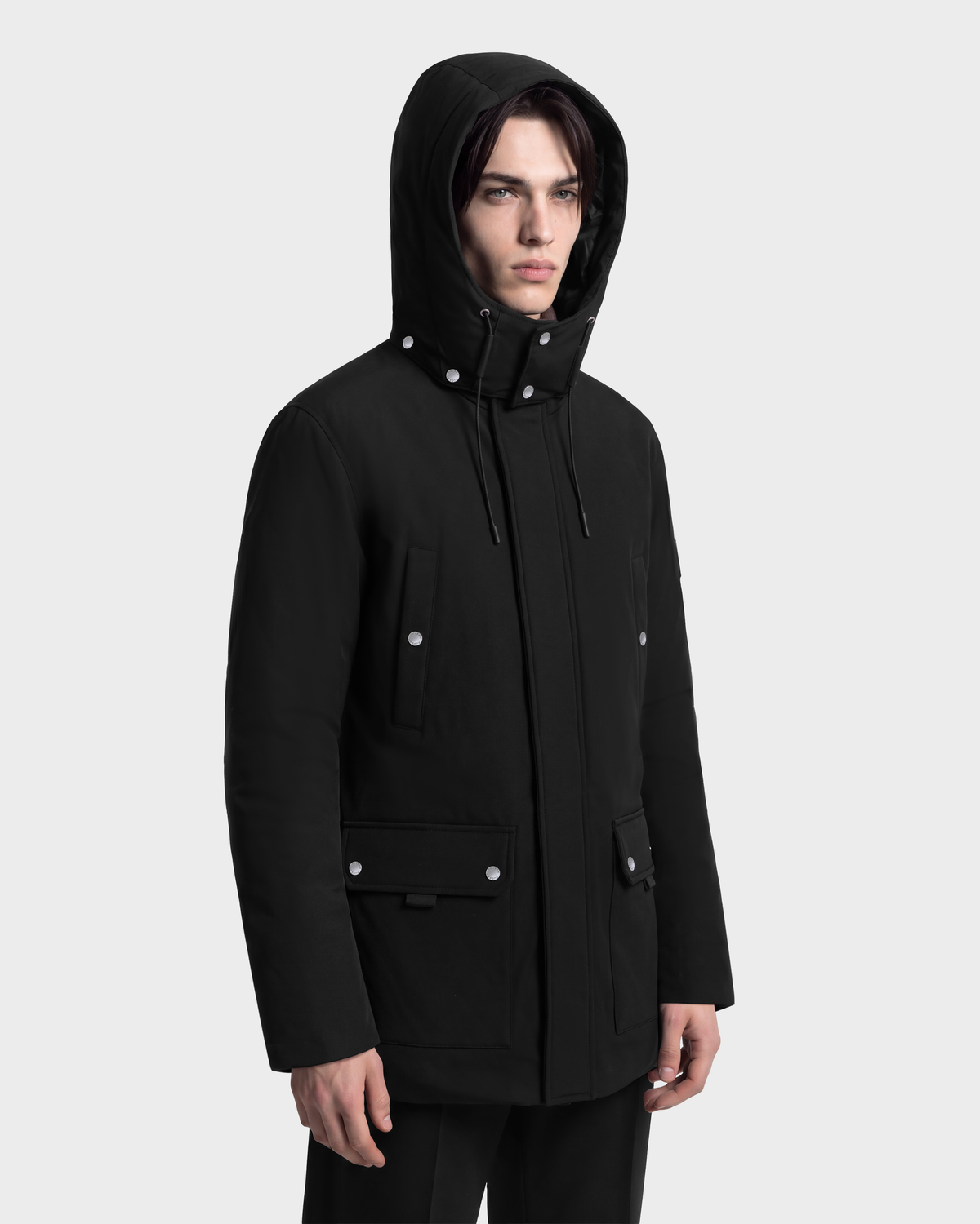 A person wears a black RUDSAK EDWARD - 6126501 Black extreme winter hooded parka with silver snap buttons and four front pockets.