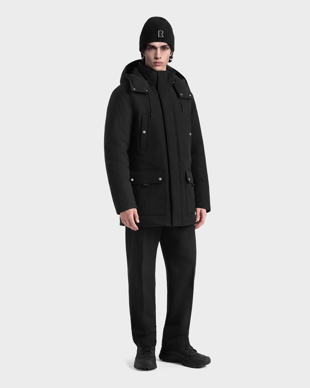 A man wearing an EDWARD - 6126501 Black Rudsak parka, black trousers, black sneakers, and a black beanie with a logo on the front.