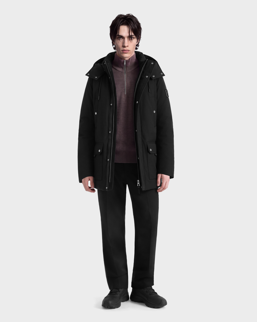 A man wearing an EDWARD - 6126501 Black deep-winter parka over a mauve zip-up sweater, black trousers, and black sneakers.