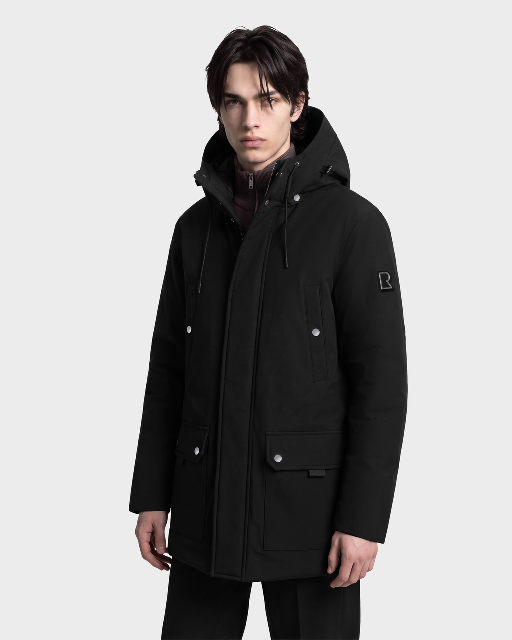 A man wearing a black RUDSAK EDWARD - 6126501 parka featuring two lower flap pockets, a button-down placket, and a small logo patch on the sleeve.