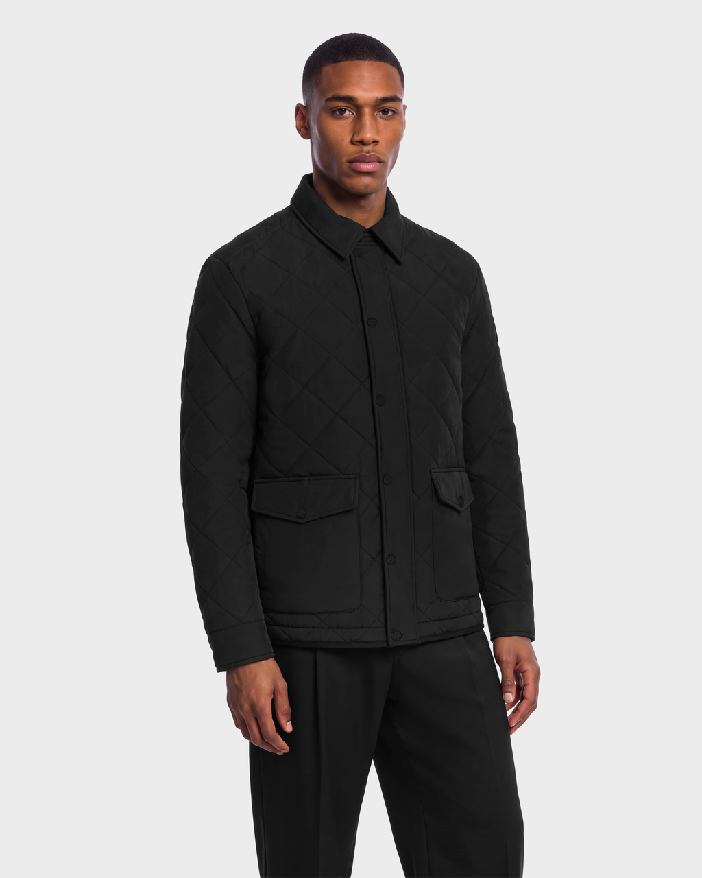 A man wearing a black Rudsak HENDRIX - 6126721 quilted collared jacket with snap closures and two front flap pockets.