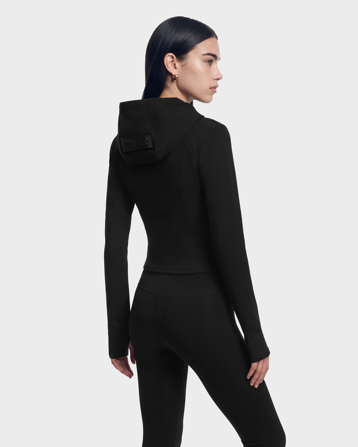 A person wearing a JUNIPER - 8526679 Black/Black fitted zip hoodie with a cropped silhouette and matching leggings.