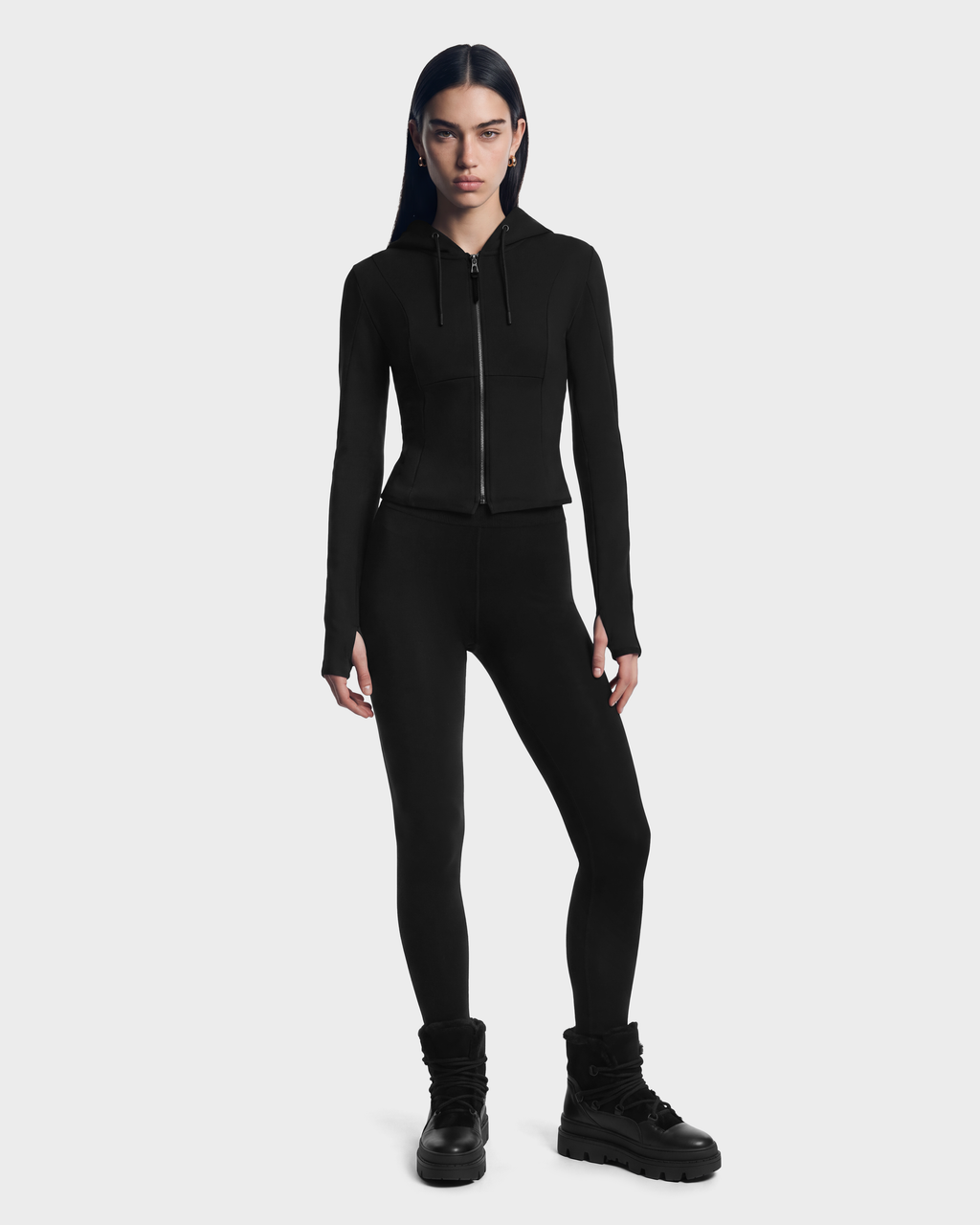 A model wearing a JUNIPER - 8526679 Black/Black fitted zip hoodie in a cropped silhouette, paired with black leggings and black lug-sole boots.
