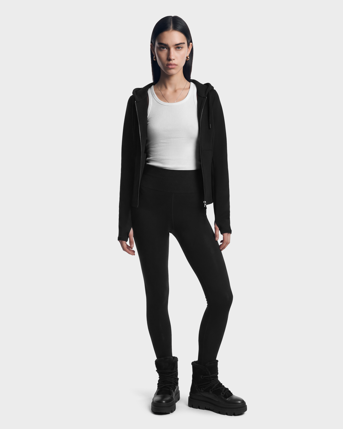 A woman wearing a JUNIPER - 8526679 Black/Black fitted zip hoodie over a white tank top, black leggings, and black boots.