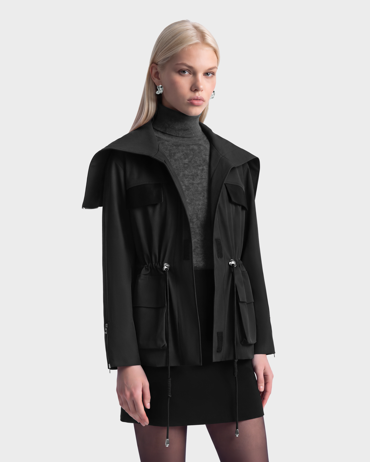 A model wearing a black ROSALINDA - 8126523 Black hooded insulated jacket with a drawstring waist and flap pockets over a grey turtleneck and black skirt.
