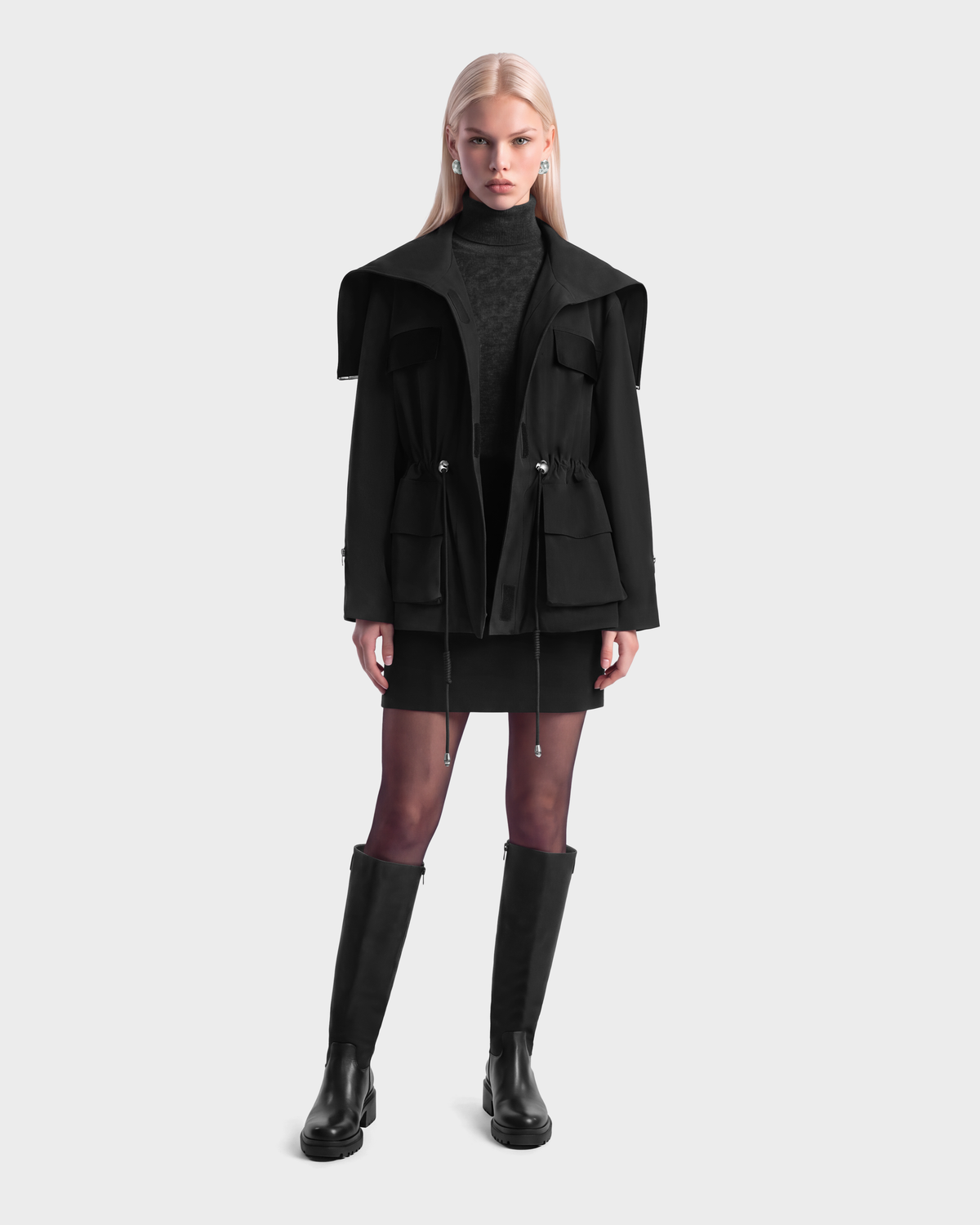 A person wearing a ROSALINDA - 8126523 Black insulated coat with a wide collar and drawstring waist, a black turtleneck, a short black skirt, and black knee-high boots.