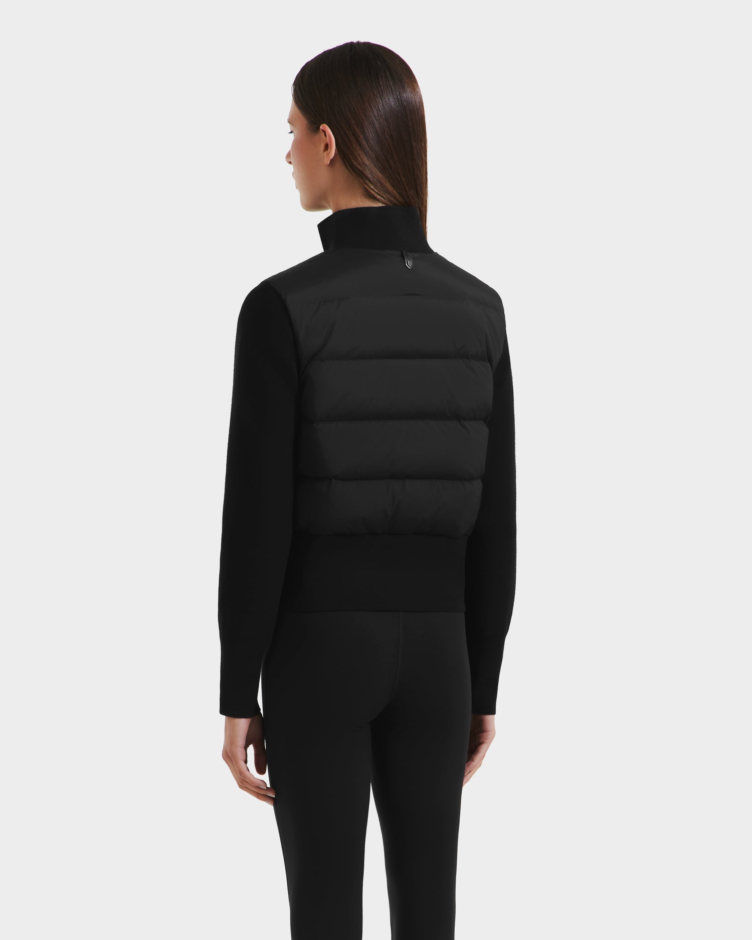 A woman with straight brown hair, wearing the Rudsak CLAREMONT - 8125502 Black short down jacket and black leggings, stands facing away from the camera against a plain light gray background.