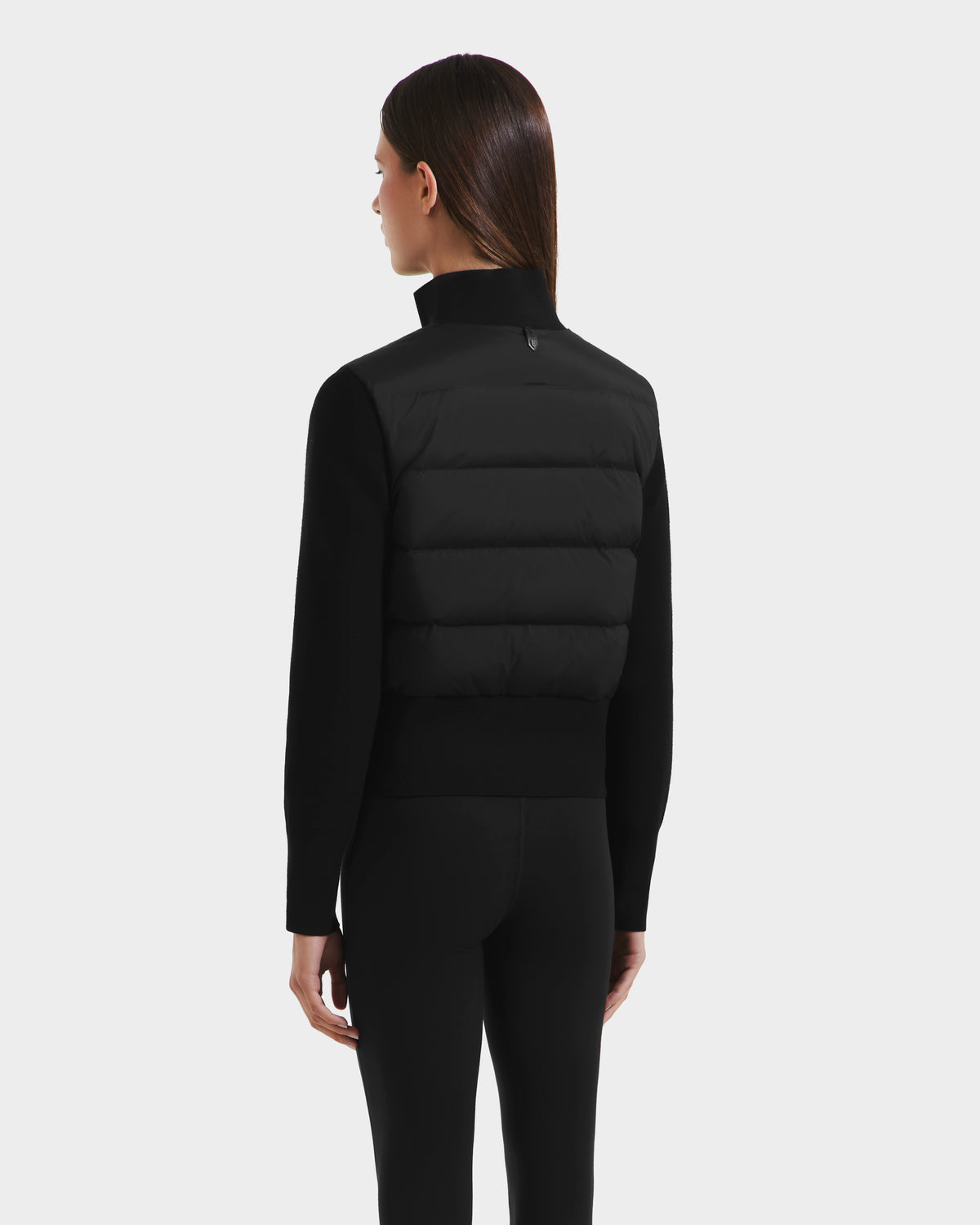 A woman with straight brown hair, wearing the Rudsak CLAREMONT - 8125502 Black short down jacket and black leggings, stands facing away from the camera against a plain light gray background.