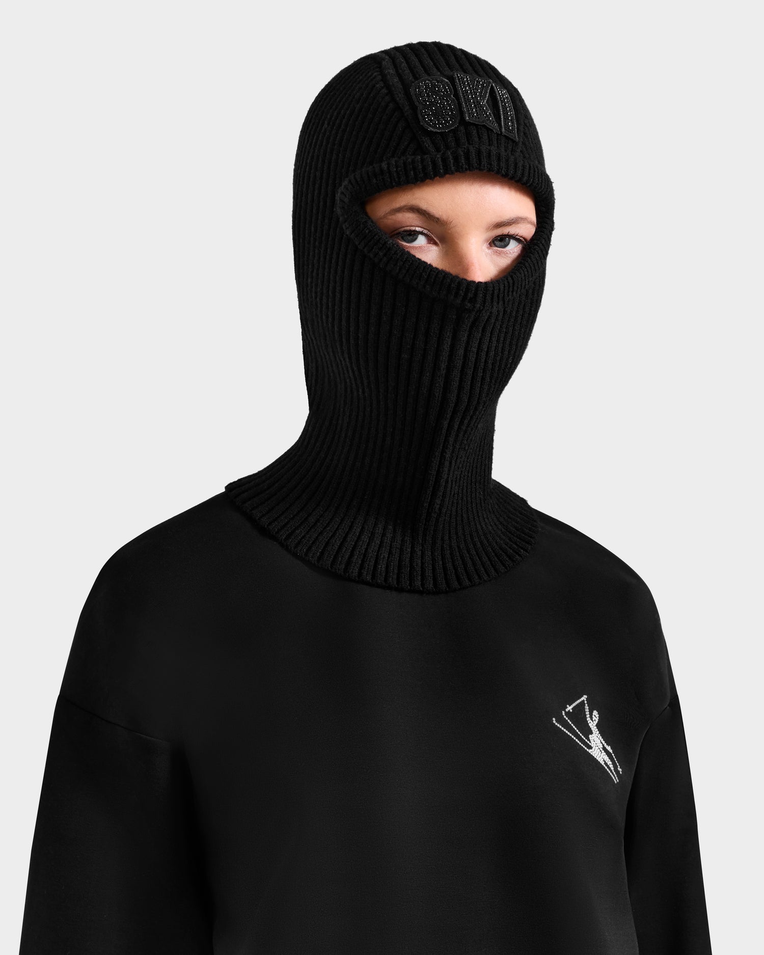 A person wears the Rudsak DIXIE - 8425551 Black women’s fitted knit balaclava with eye and mouth openings and a black sweatshirt with a crystal-embellished SKI logo, set against a plain light background—a chic winter look.