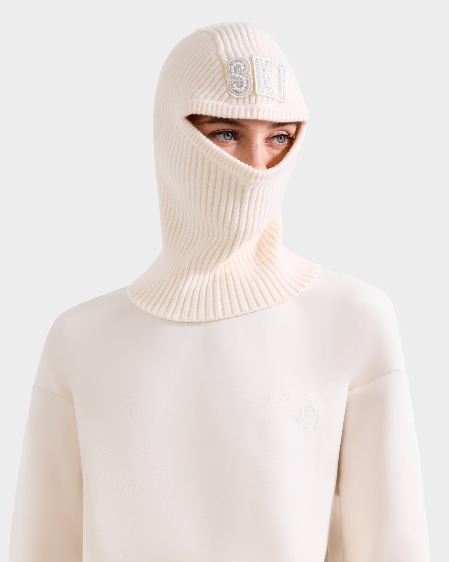 A person wears the Rudsak DIXIE - 8425551 Vanilla women’s fitted knit balaclava with a crystal-embellished SKI logo, paired with a matching cream sweater, set against a light gray background—an elegant winter wardrobe addition.