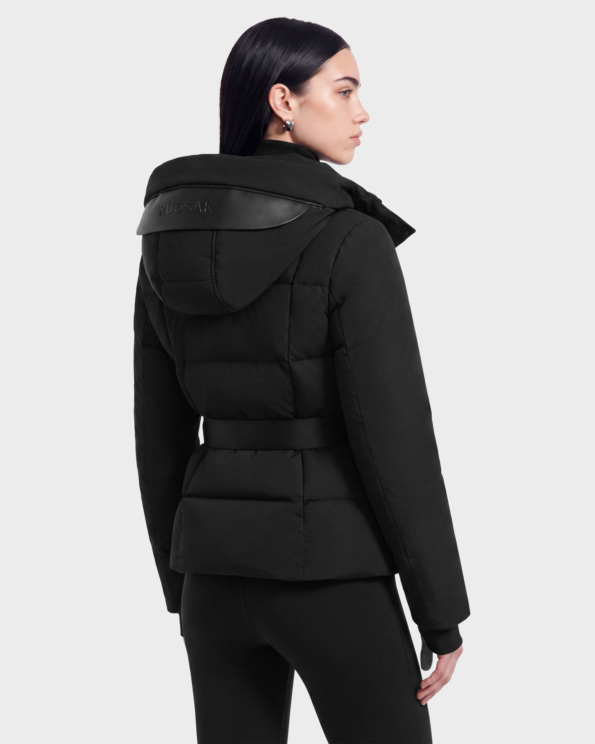 A woman with straight dark hair wears the ADELINE - 8124544 Black women’s down puffer jacket by Rudsak, styled with black pants. She stands against a light gray background, facing away and turning her head slightly to the side.