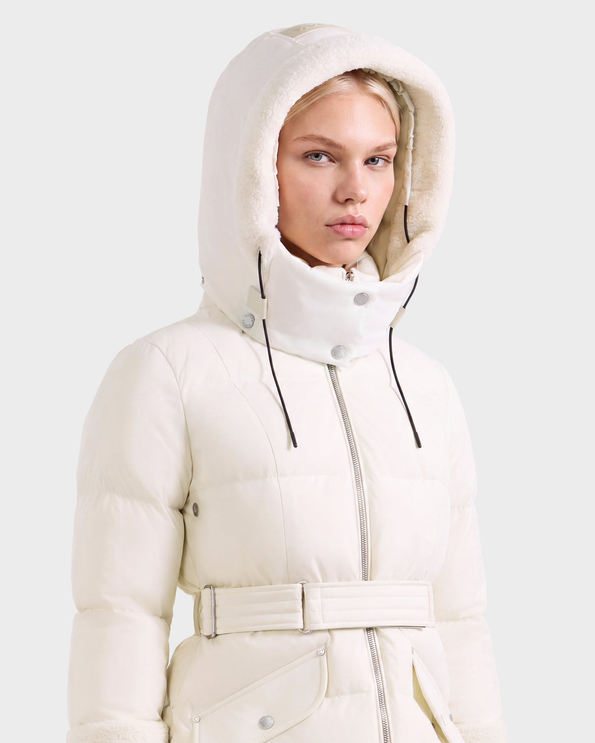 A woman wears a vanilla-colored AUDREY L - 8124585 Rudsak knee-length down puffer jacket with a belted waist and a fleece-lined hood.