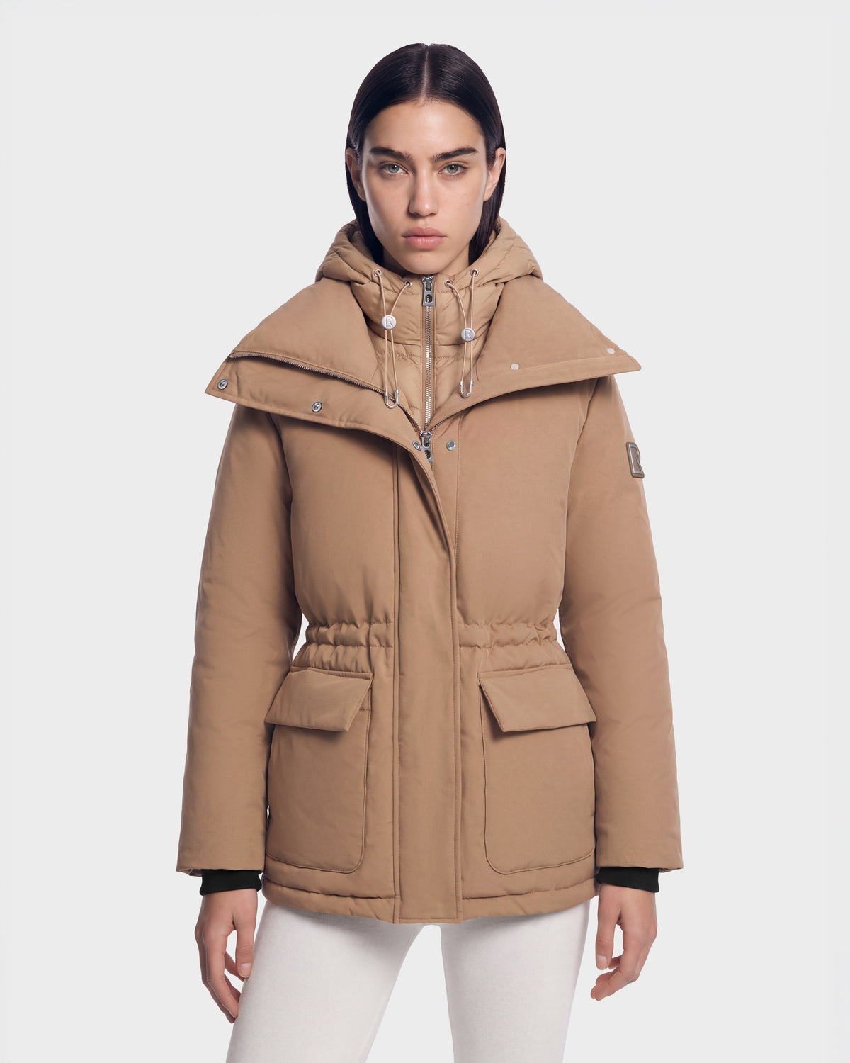 A person wears a BHIMA - 8126515 Camel hooded puffer jacket in camel, featuring a wide collar, drawstring waist, and two front flap pockets.