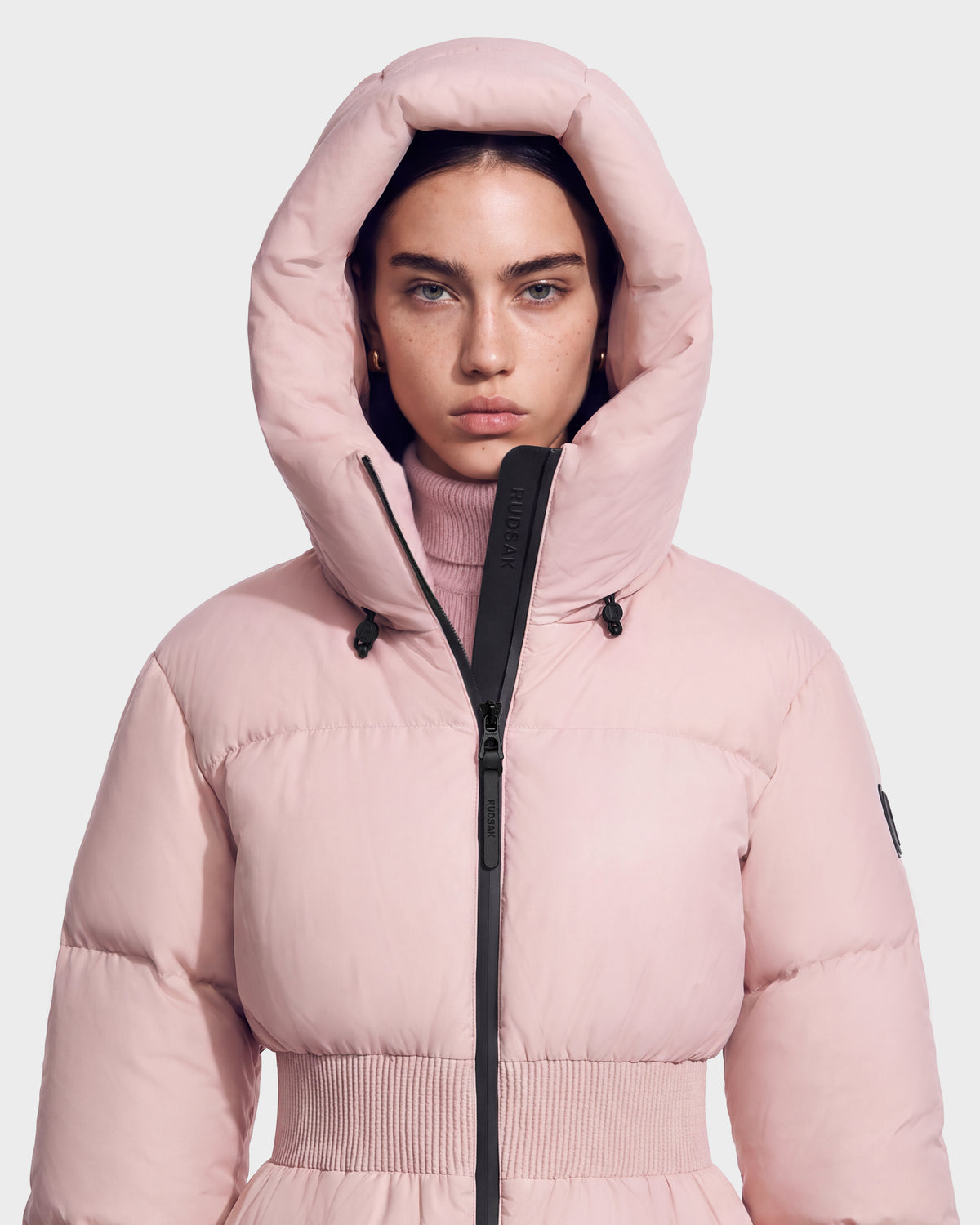 A person wearing a pale pink Rudsak DIAMOND - 8126772 Vintage Rose hooded quilted puffer jacket with a cinched waist.