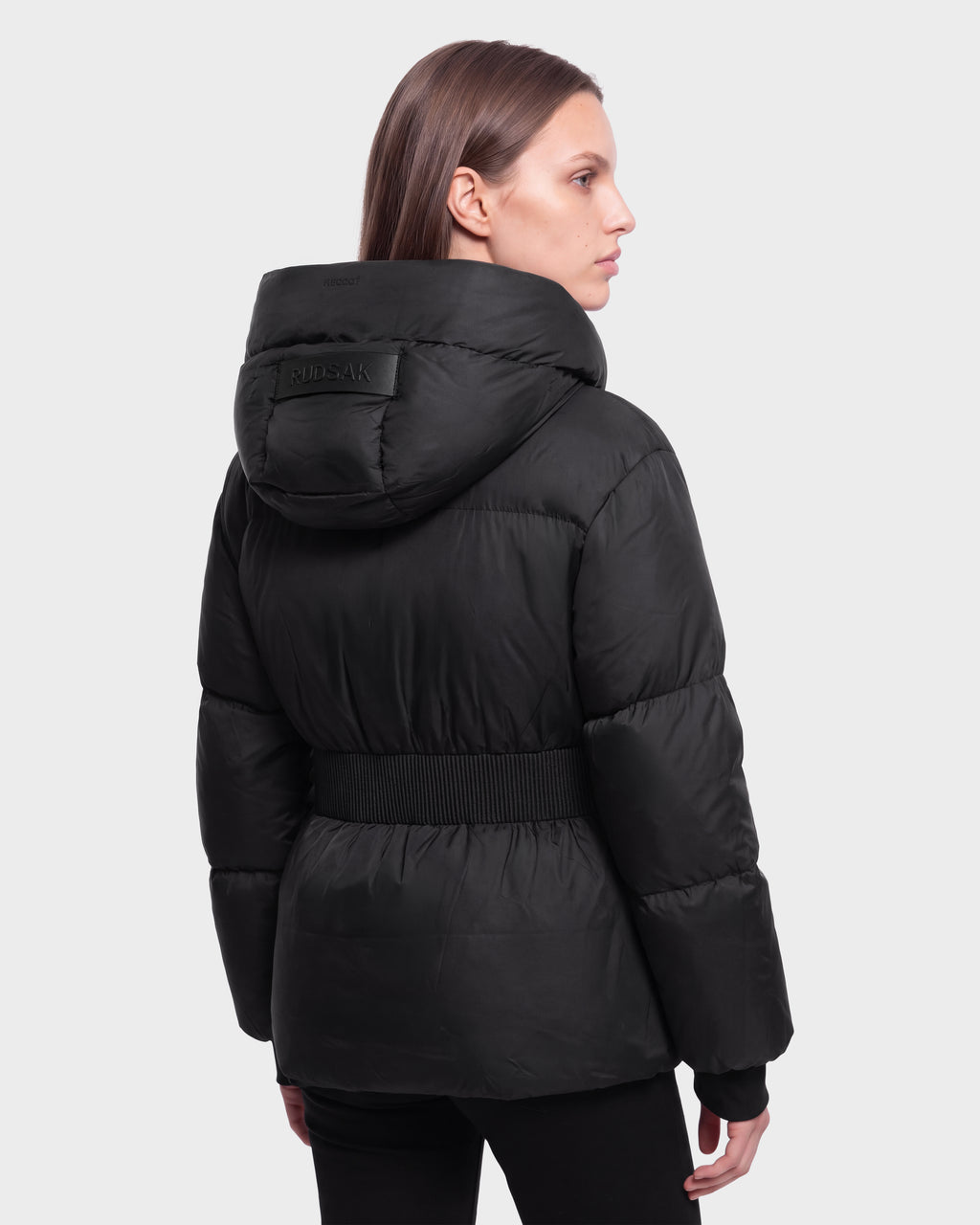 Woman wearing the Rudsak DIAMOND - 8126772 Black puffer jacket with hood, seen from behind at an angle against a plain light gray background.
