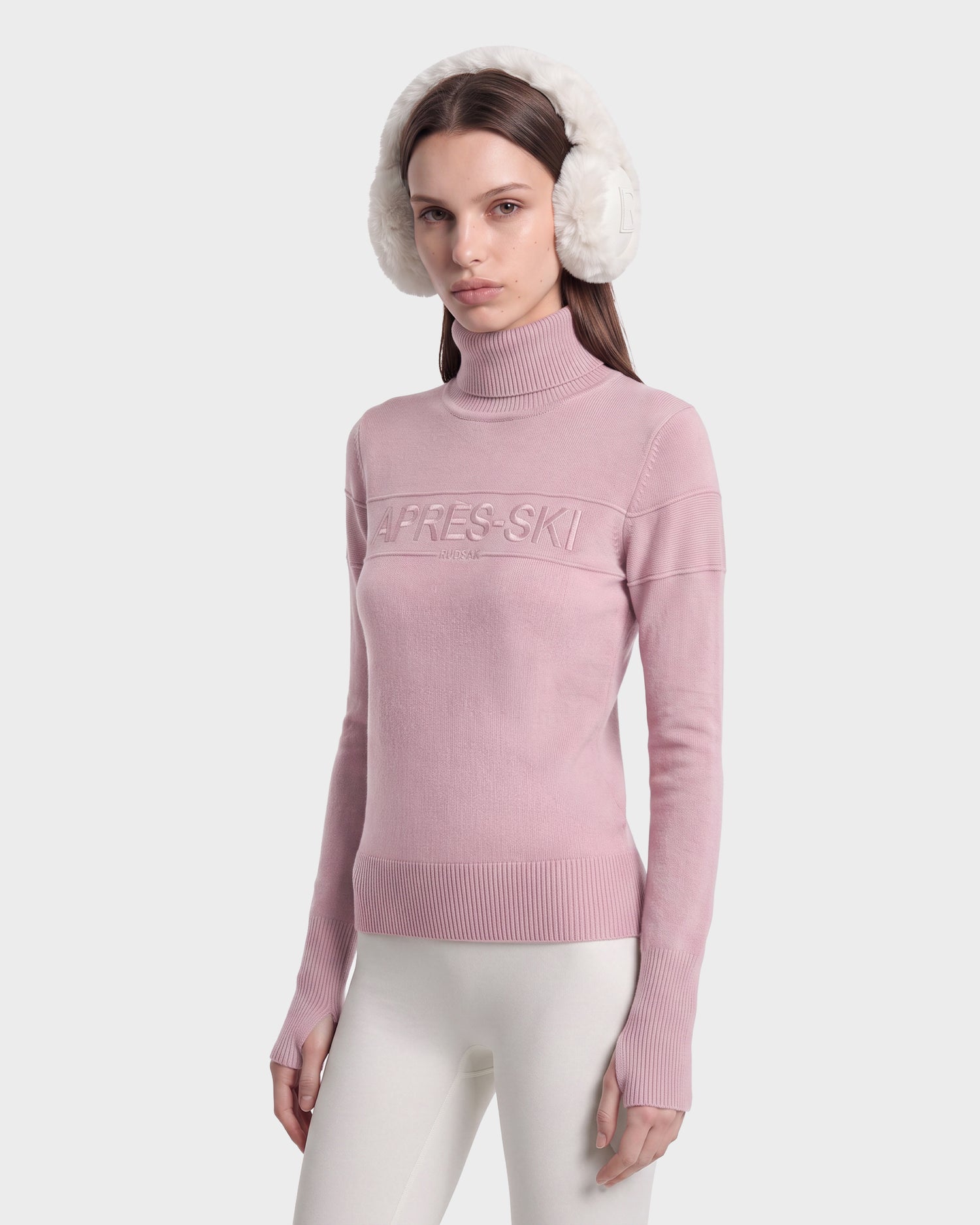Wearing the Rudsak ERIKA - 8525705 Vintage Rose apres-ski sweater with “APRES-SKI” embossed on the chest, paired with white earmuffs and white leggings, a woman stands against a plain white background—a cozy look that’s perfect for any winter wardrobe.