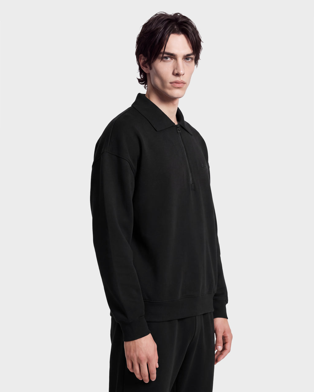 A person with medium-length dark hair wears the Rudsak IDRIS - 6526500 Black polo collar sweater and matching pants, standing against a plain white background, looking slightly to the side with a neutral expression.