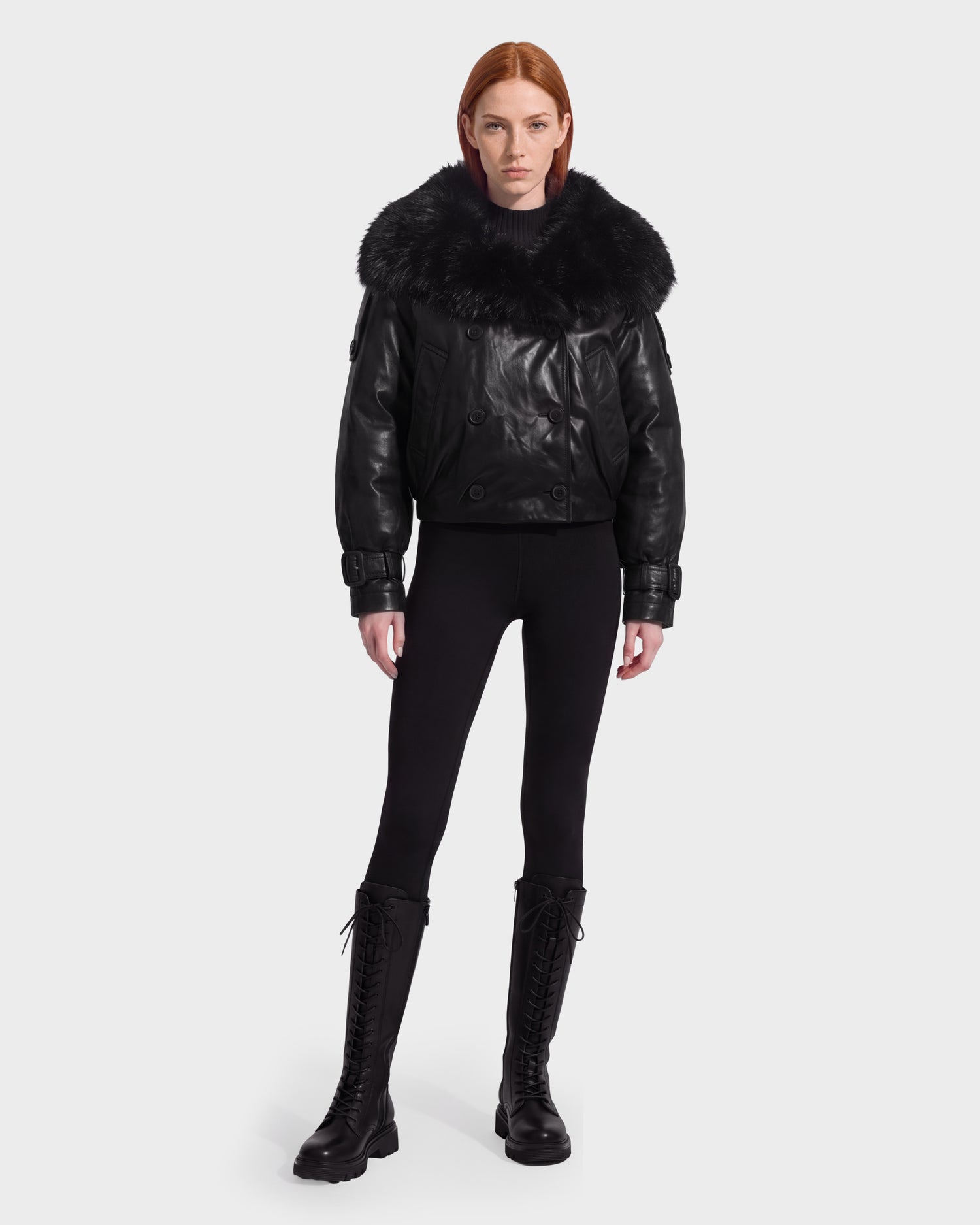 A person with straight red hair wears the Rudsak JANELLE - 8126936 Black leather bomber jacket with a large faux fur collar, paired with black leggings and black lace-up boots, standing against a plain white background.