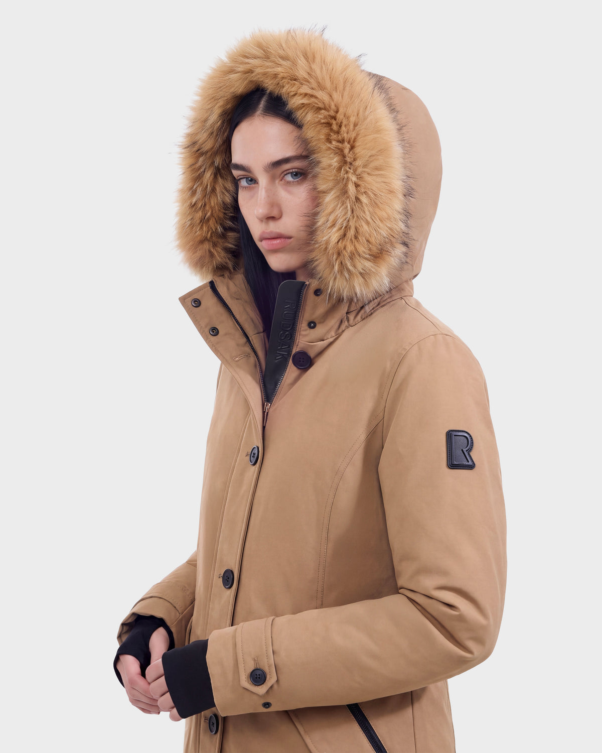 A person wears the KEYSHIA - 8126517 Camel by Rudsak, a tan insulated jacket with a fur-lined hood and a black R patch on the sleeve. They face the camera against a plain white background.