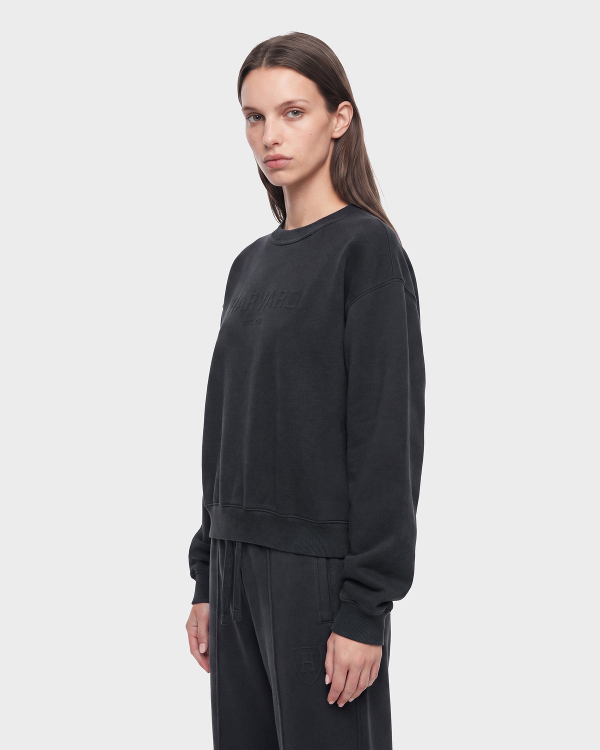 A person with long brown hair, dressed in the Rudsak LEXYE HV - H526900 Titanium loose-fitting black sweatshirt and matching pants, stands against a plain light gray background, facing slightly to the left with a neutral expression.