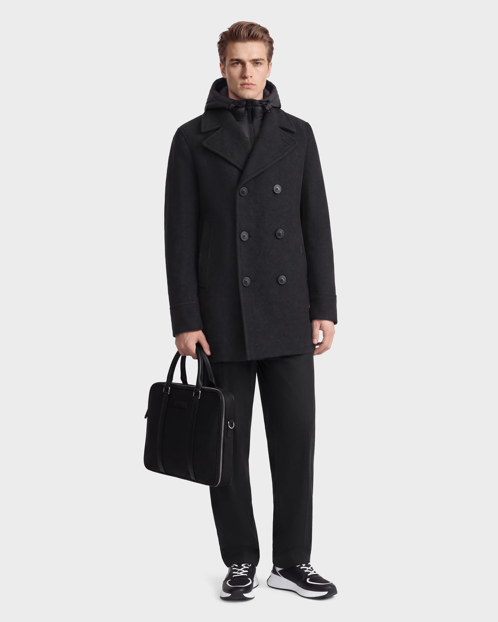 A man wearing a charcoal LUCIEN - 6126508 double-breasted wool-blend pea coat over a black hooded jacket, dark trousers, and black-and-white sneakers, holding a black briefcase.
