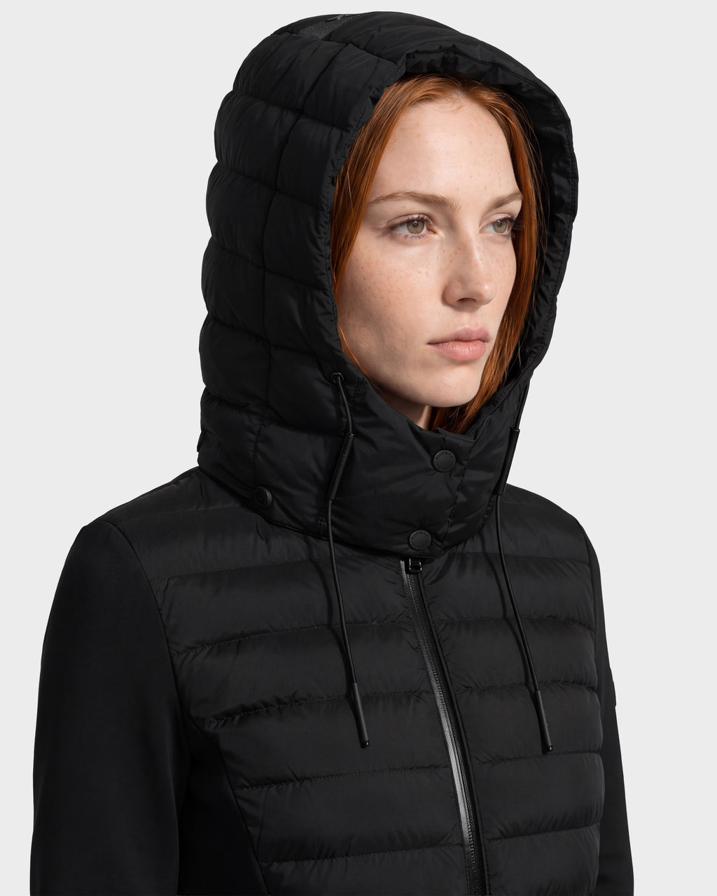 A woman with straight red hair models the Rudsak MERINA - 8125056 Black hooded quilted down jacket, gazing slightly to the side against a plain light background.