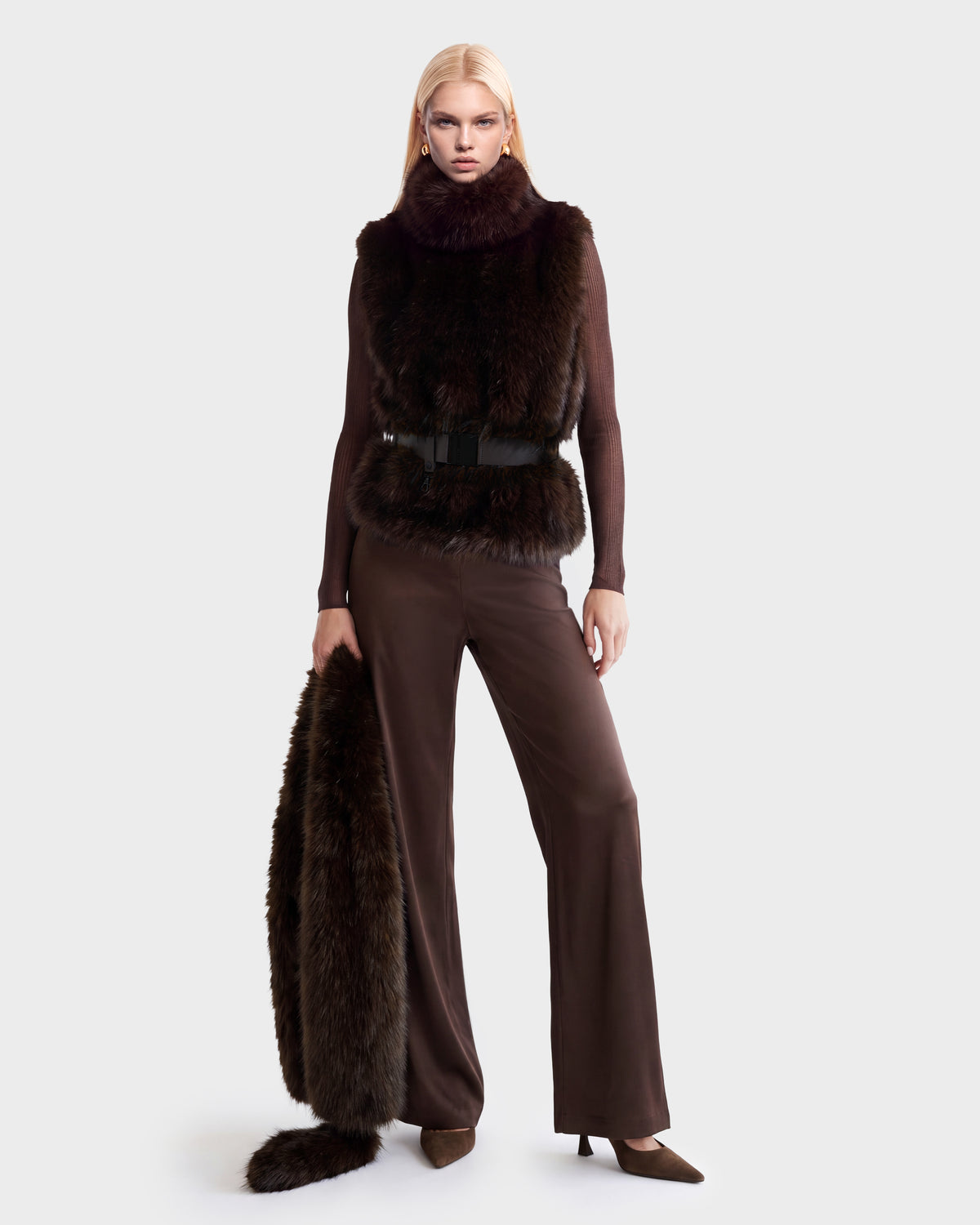 A model wears the Rudsak MAULIN - 8524611 Mix Espresso brown mock-neck pullover, matching trousers, and a dark brown faux fur vest cinched with a black belt, while holding a matching fur garment.
