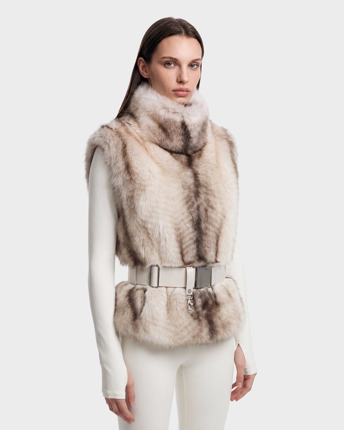 A woman wearing a Rudsak MAULIN - 8524611 Mix Vanilla faux fur vest in beige and brown with a high neck and buckled waist belt over a white mock-neck pullover.